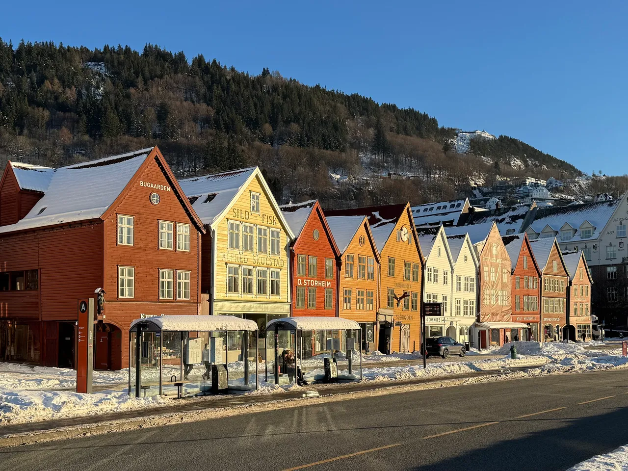 A row of steep-gabled wooden houses painted red, yellow, orange and white along the harbour front at Bryggen, with snow on the roofs and a wooded hill behind