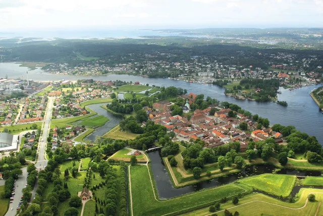 Aerial view of Fredrikstad's Old Town, its red-roofed houses and church tower enclosed by green ramparts and water-filled moats, with the wide Glomma river and the islands of the outer Oslofjord beyond