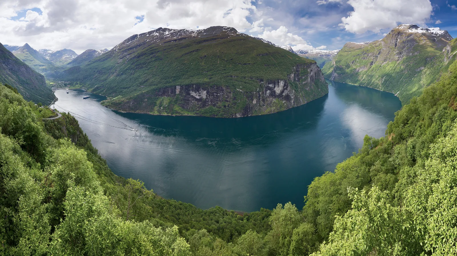 Geirangerfjord seen from high above, a deep blue fjord curving between steep green mountainsides with snow on the peaks
