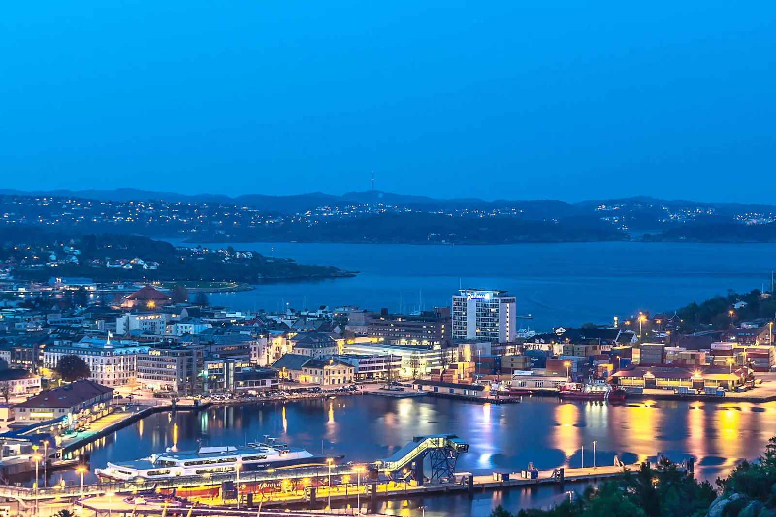 Kristiansand at dusk seen from a hillside, with lit-up buildings along the harbour, a passenger boat at the quay in the foreground and the wide fjord and wooded islands beyond under a deep blue sky