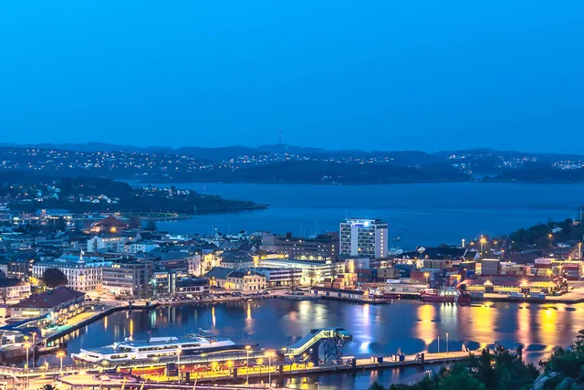 Kristiansand at dusk seen from a hillside, with lit-up buildings along the harbour, a passenger boat at the quay in the foreground and the wide fjord and wooded islands beyond under a deep blue sky