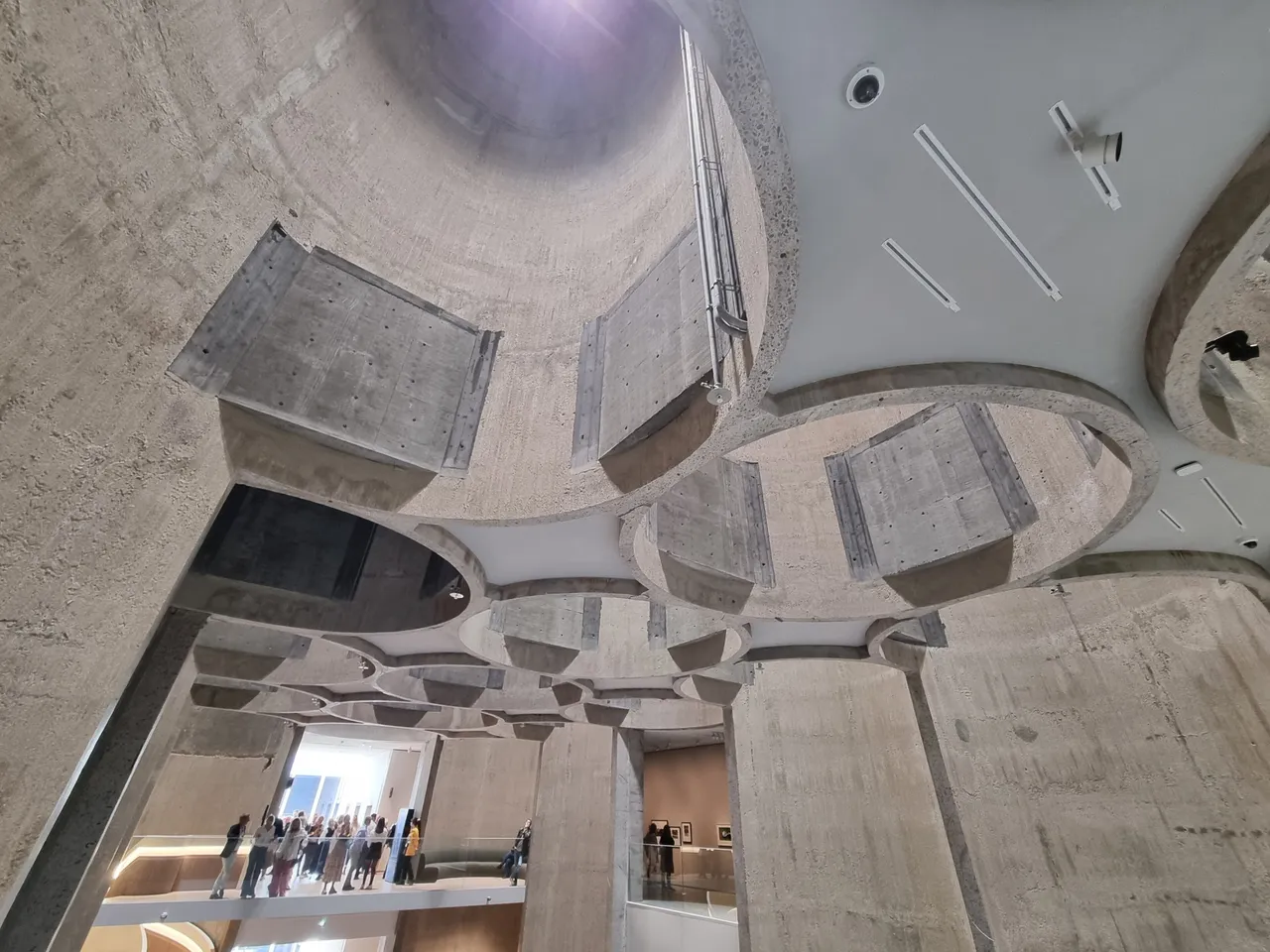 Looking up inside Kunstsilo at the cut-open concrete silo tubes forming the ceiling, with visitors on a gallery level below