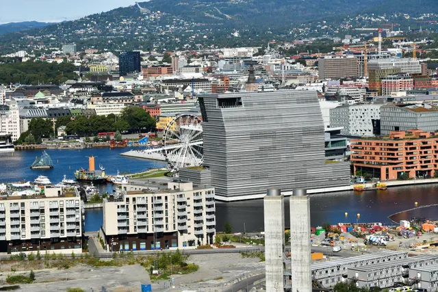 Central Oslo seen from the Ekeberg hillside on a summer day, with the tall grey MUNCH museum on the harbour in Bjørvika, the white Opera House roof and a Ferris wheel beside it, the city centre behind and the Holmenkollen ski jump on the wooded ridge above