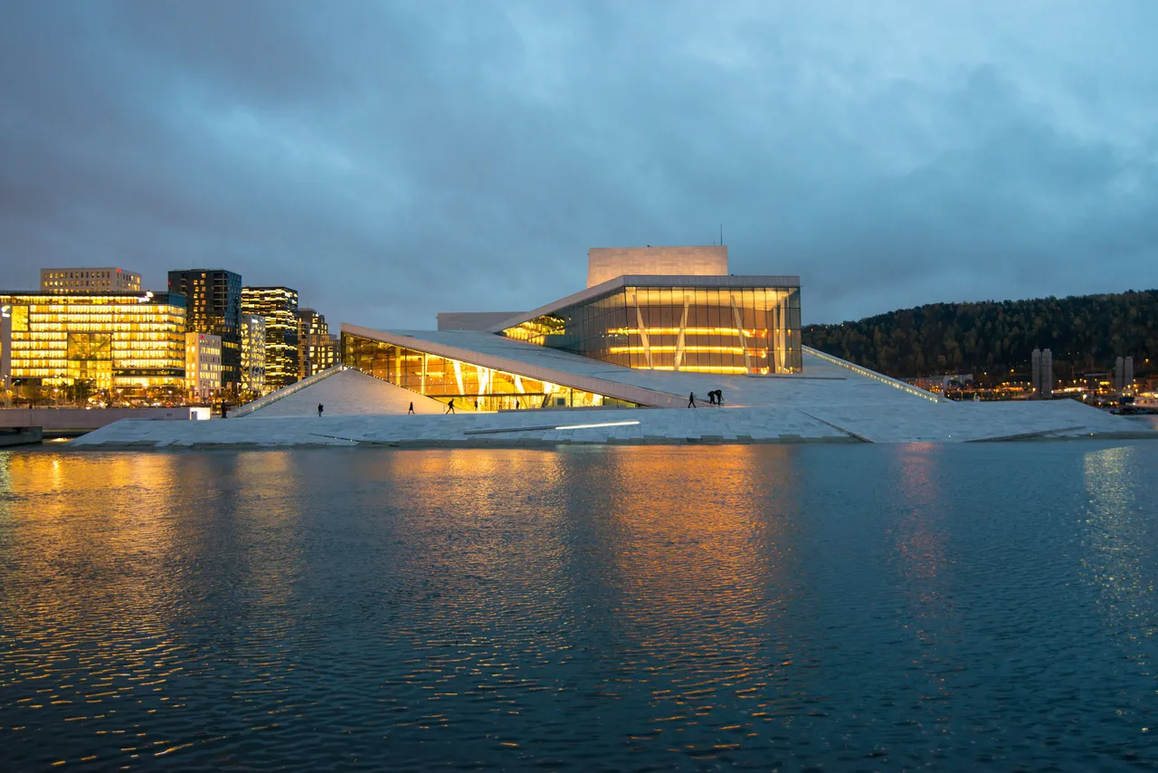 The Oslo Opera House at dusk, its sloping white roof rising from the water with a few people walking up it, the glass foyer lit golden and lit office blocks to the left
