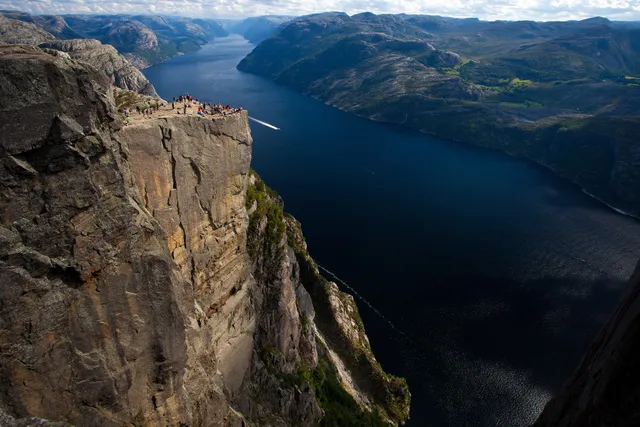 The flat top of Preikestolen crowded with hikers, its sheer grey rock face dropping straight down to the dark blue Lysefjord, with a speedboat's wake far below and green mountains stretching into the distance