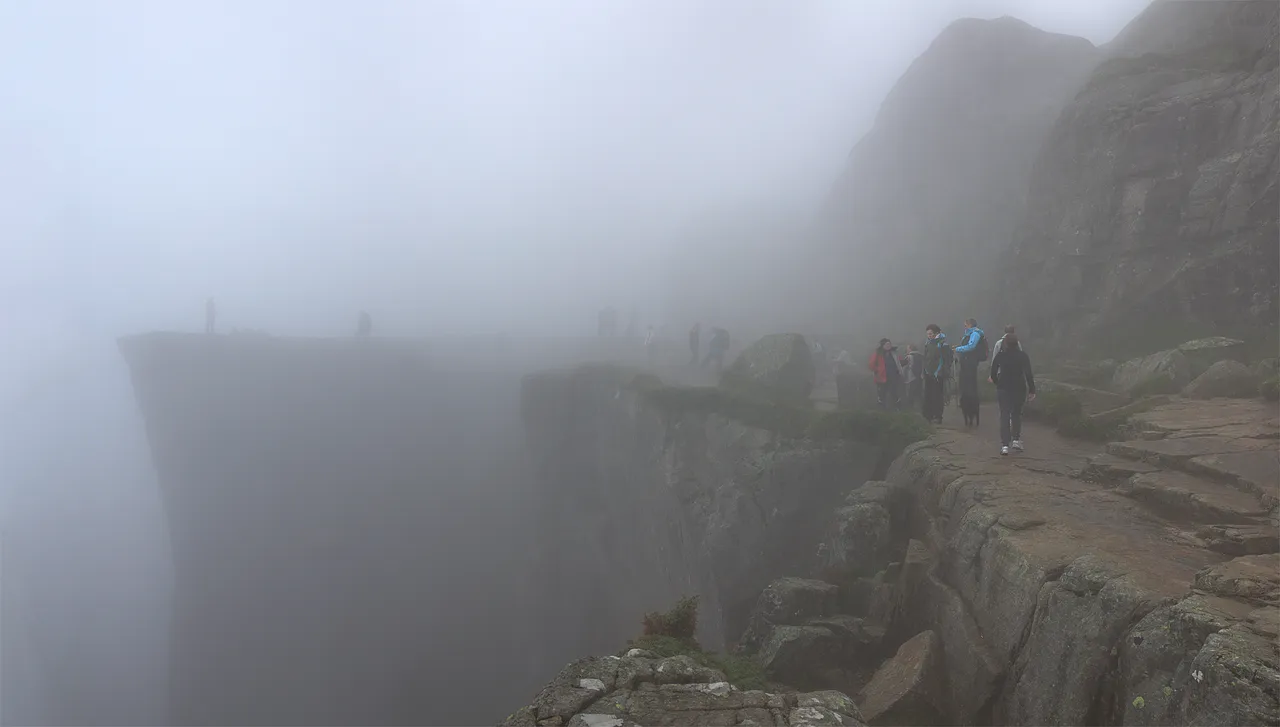 Hikers on a rocky ledge beside the flat-topped cliff of Preikestolen, its edge dropping away into thick fog