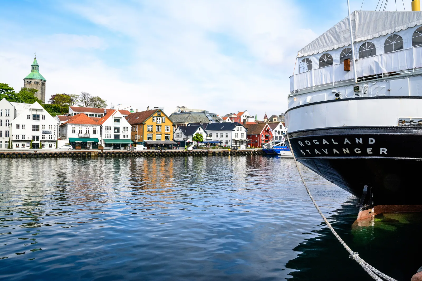 White and ochre wooden houses with restaurants along the quay of Stavanger's Vågen harbour, a stone watchtower with a green copper roof on the hill behind, and the black bow of a moored ship named Rogaland in the foreground