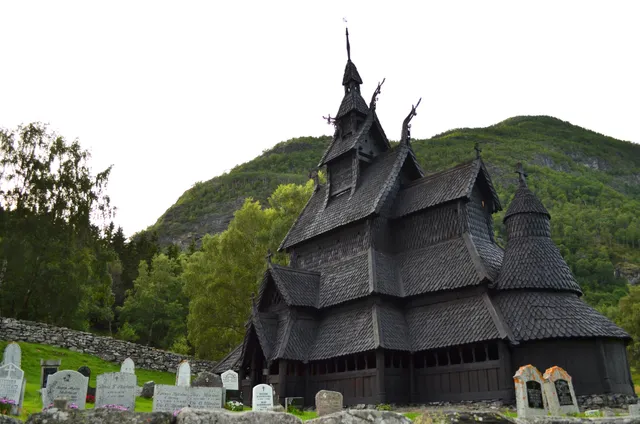 Borgund Stave Church seen from the churchyard, a dark wooden church with tier upon tier of shingled roofs, carved dragon heads on the gables and a slender spire, below a green wooded mountainside