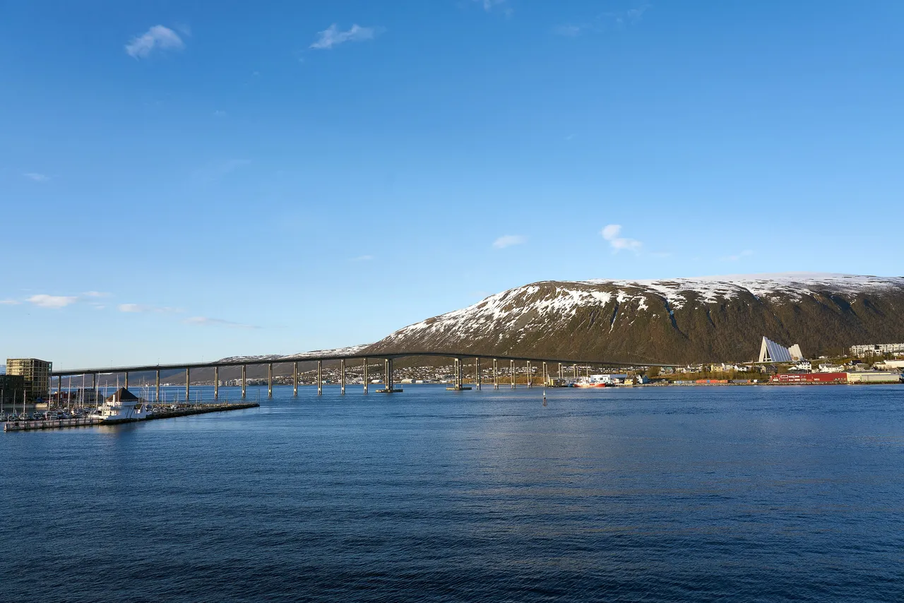 The long Tromsø Bridge crossing a deep blue sound to the white, triangular Arctic Cathedral at the foot of a snow-streaked mountain under a clear blue sky
