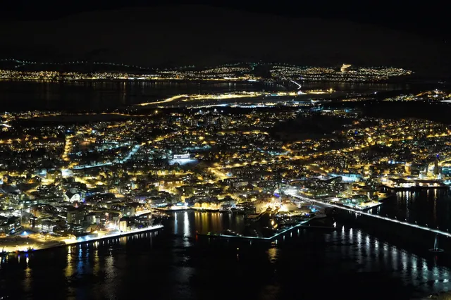 Tromsø at night seen from the mountain above the city, its lit streets and harbour reflected in the sound, the long bridge on the right and the airport runway lit up behind the town