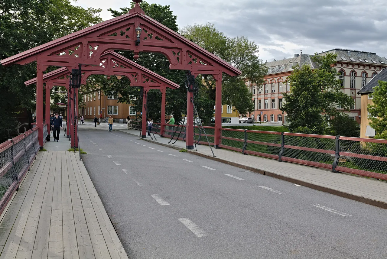 The Old Town Bridge in Trondheim, with its two dark red carved wooden gate portals, plank walkways on either side and pedestrians crossing towards wooden and brick buildings