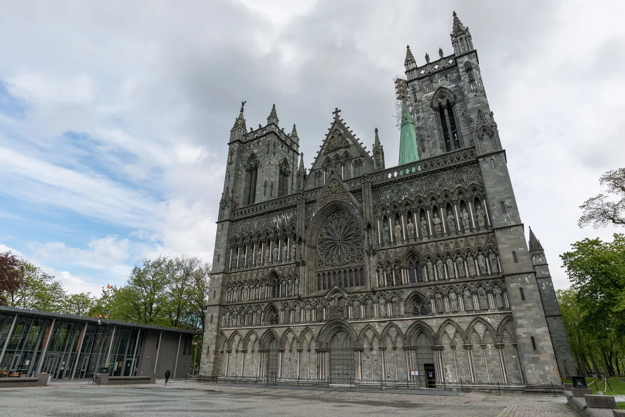 The grey soapstone west front of Nidaros Cathedral, with two towers, a rose window and rows of carved statues, beside the low glass visitor centre under a cloudy sky